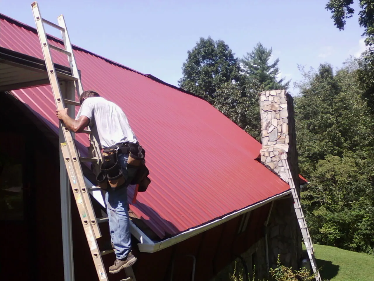 Roofer on a ladder beside a red metal roof and stone chimney — Soffit & Fascia Repair by Roofing Brothers in Spring Branch